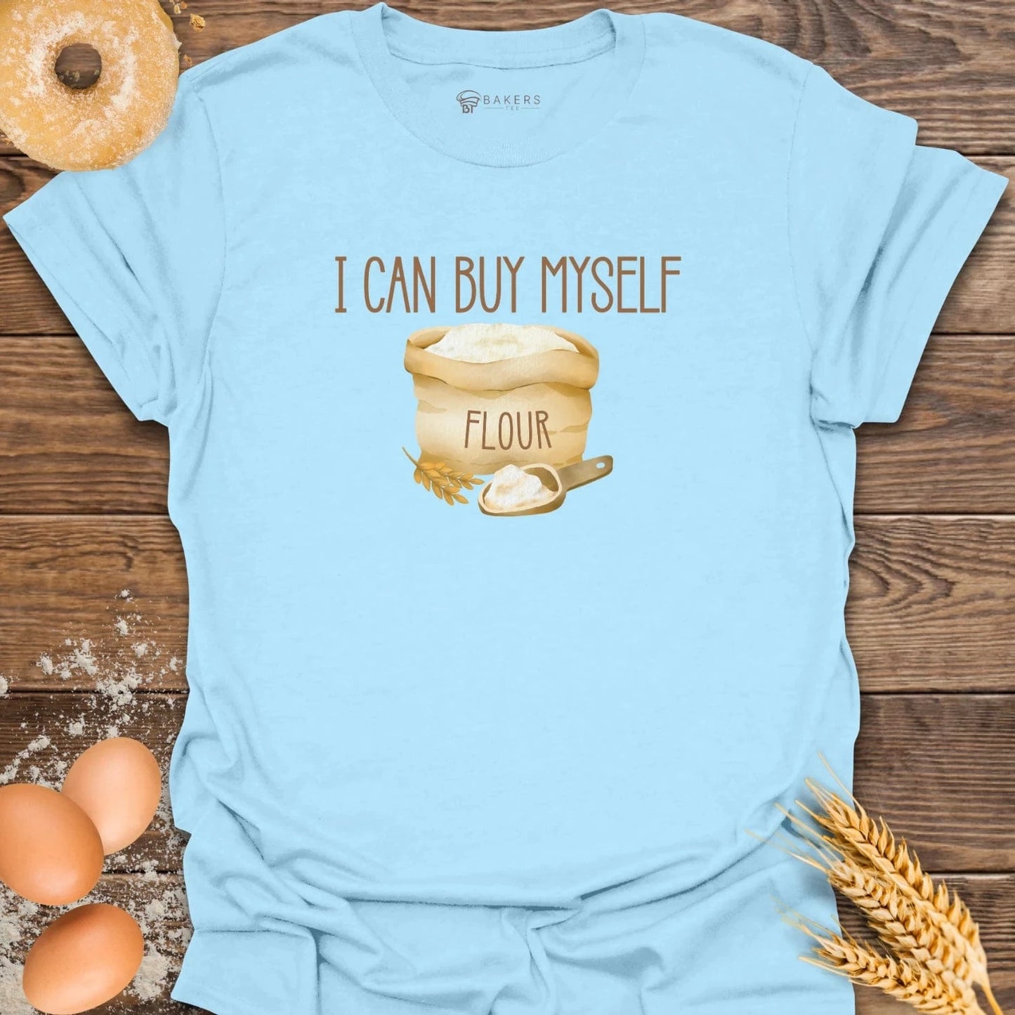 A Light Blue crew neck t-shirt with the text 'I CAN BUY MYSELF FLOUR' printed on the front, presented on a wooden background surrounded by baking ingredients like flour and wheat. By Bakers Tee