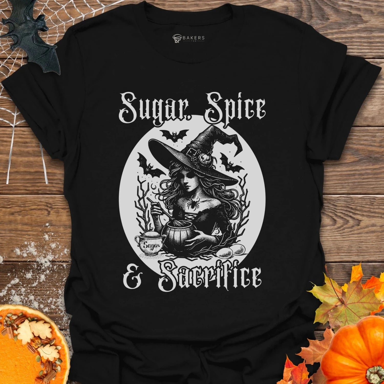 Black t-shirt with a witch graphic and 'Sugar, Spice & Sacrifice' text on a wooden surface with pumpkins and leaves.