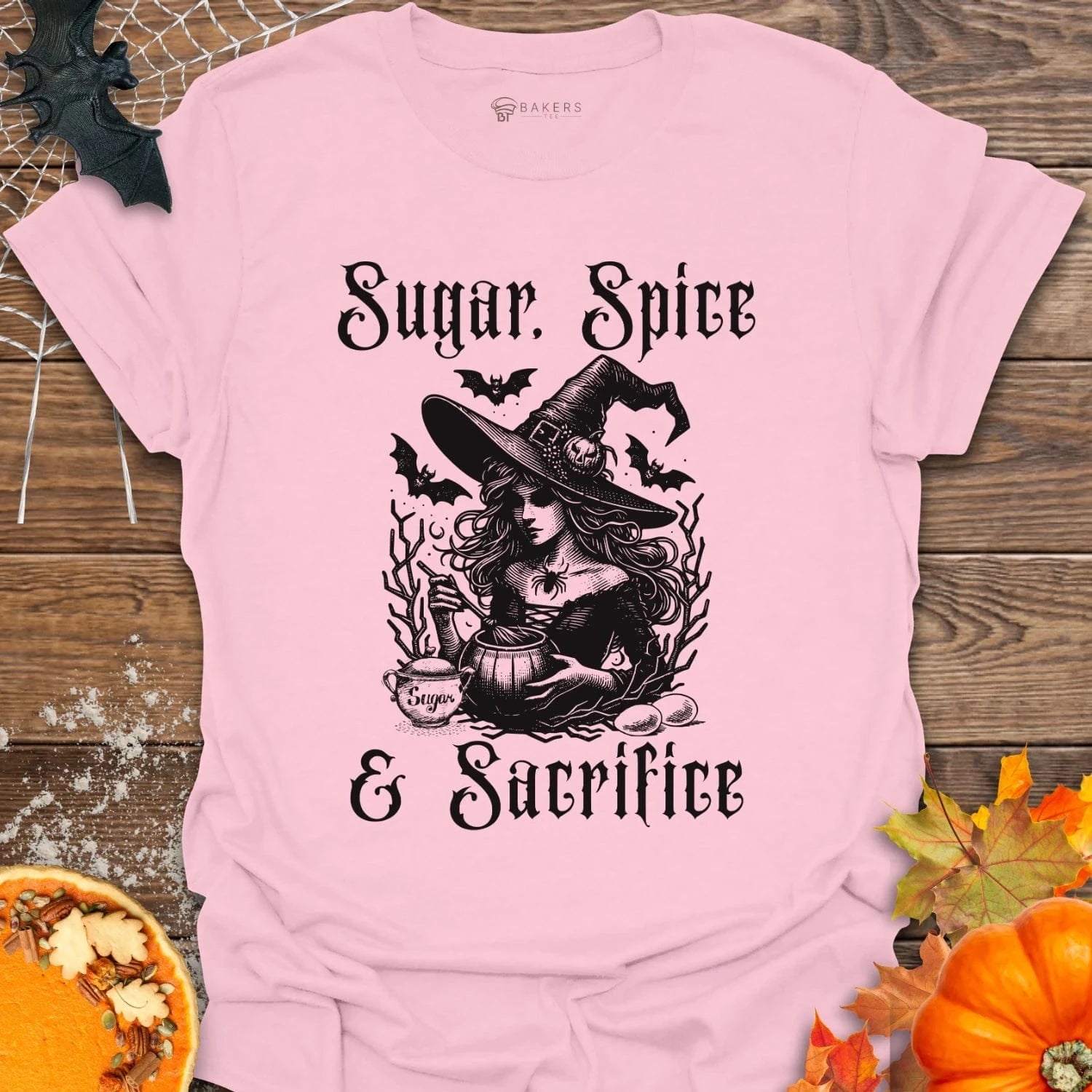 Pink t-shirt with Halloween-themed design featuring a witch and text on a wooden background with pumpkins and leaves.