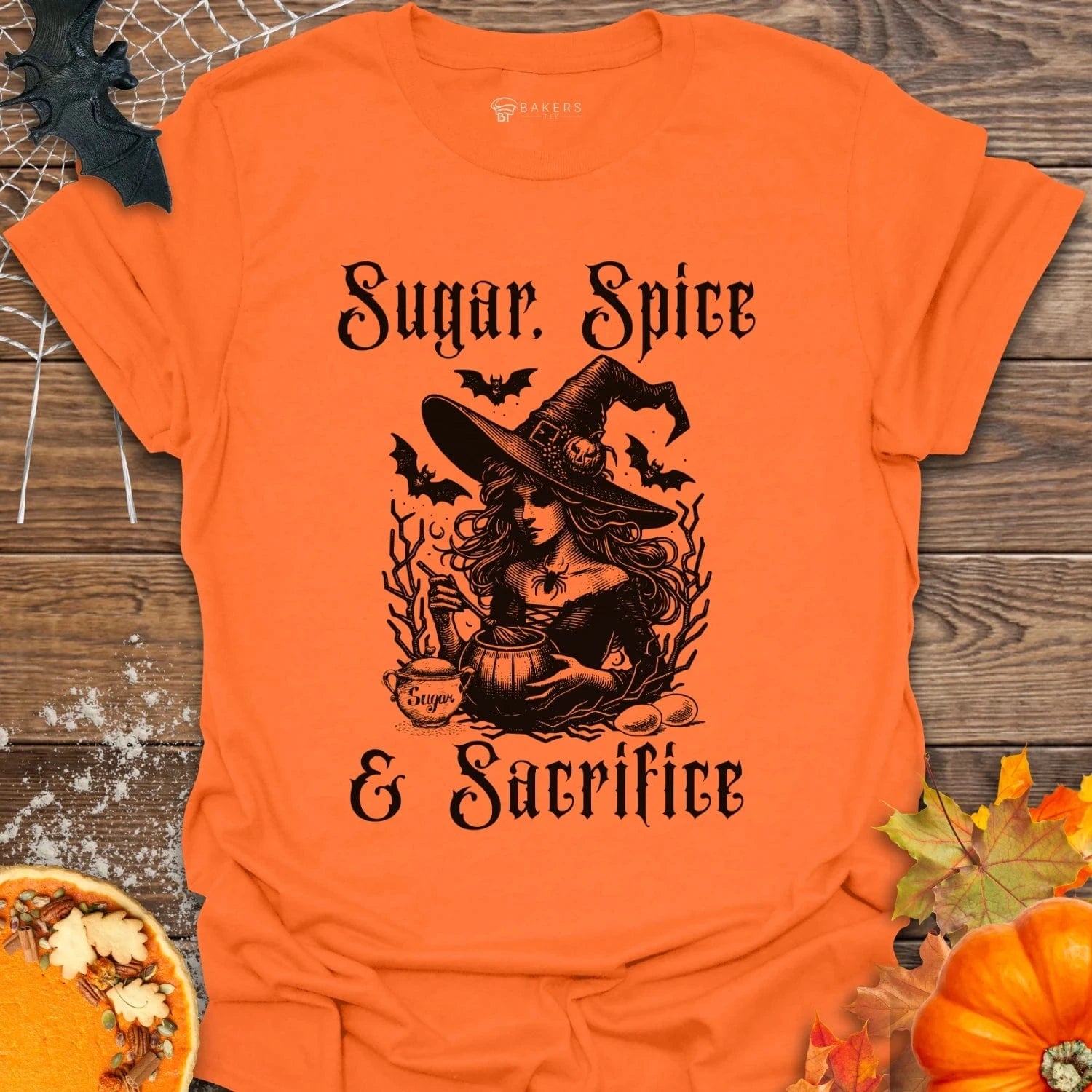 Orange t-shirt with witch graphic and text 'Sugar, Spice & Sacrifice' on a wooden surface with pumpkins and leaves.