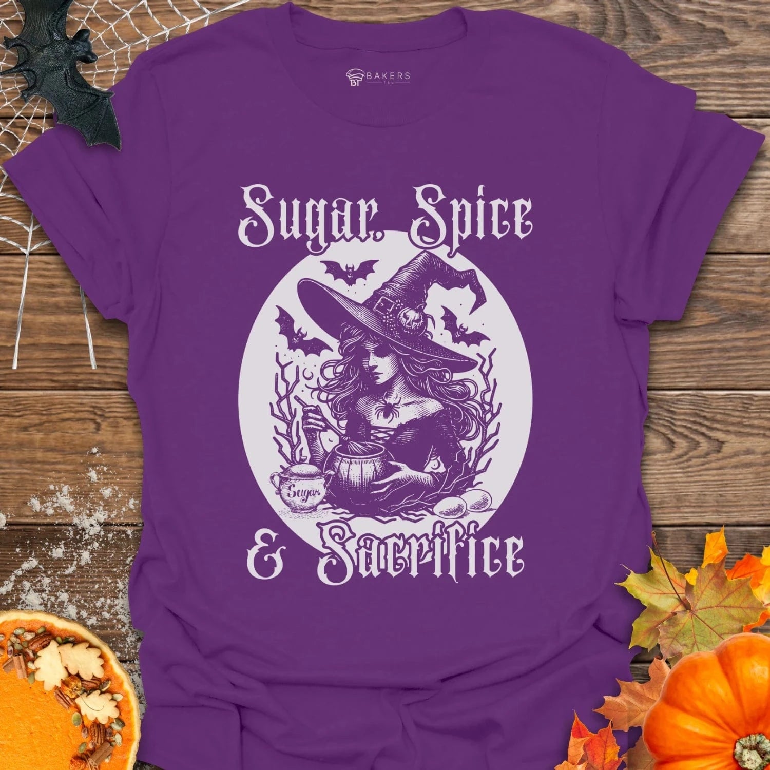 Purple t-shirt with witch graphic and 'Sugar, Spice & Sacrifice' text on a wooden surface with pumpkins and leaves.