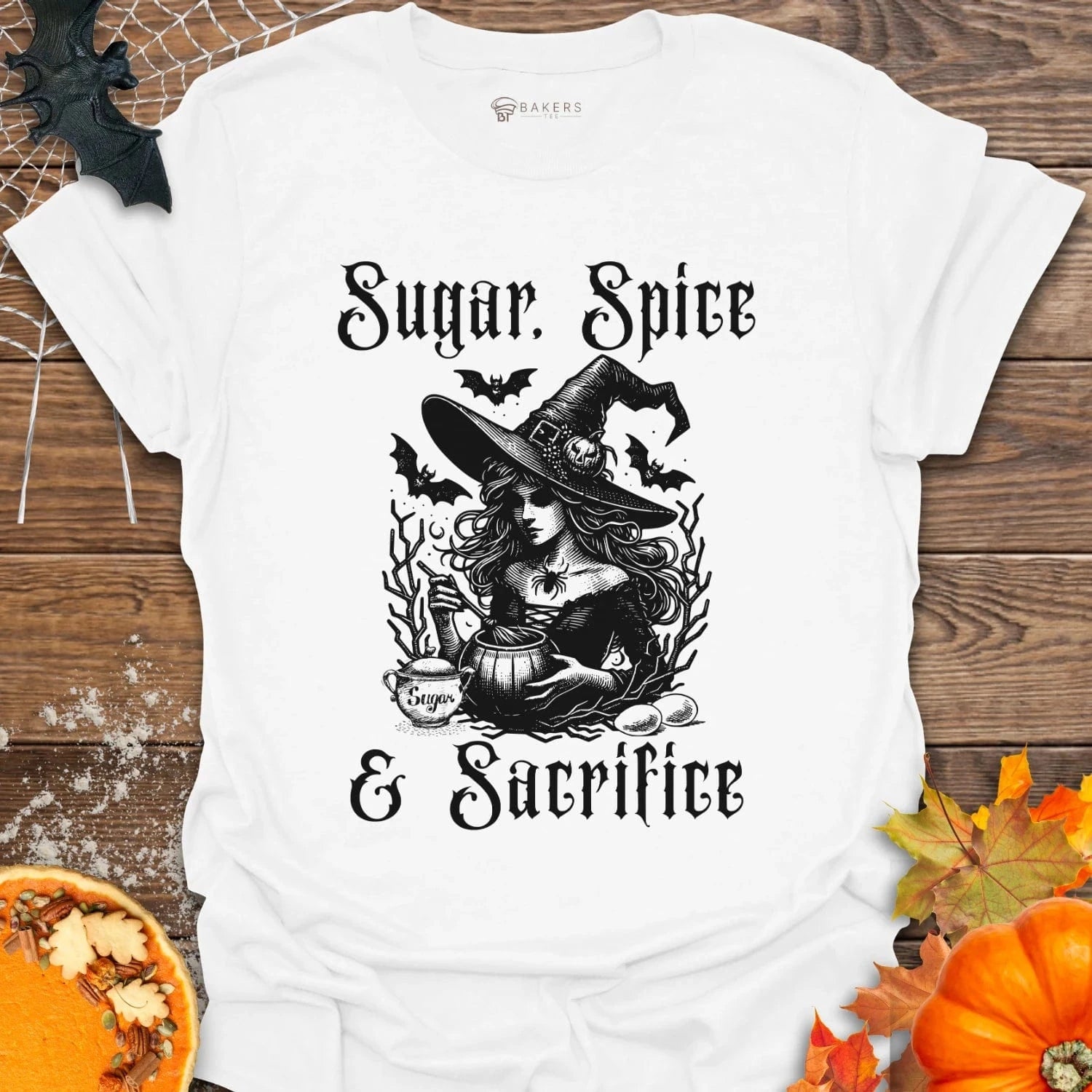 White t-shirt with Halloween-themed design featuring a witch and text on a wooden surface with pumpkins and leaves.