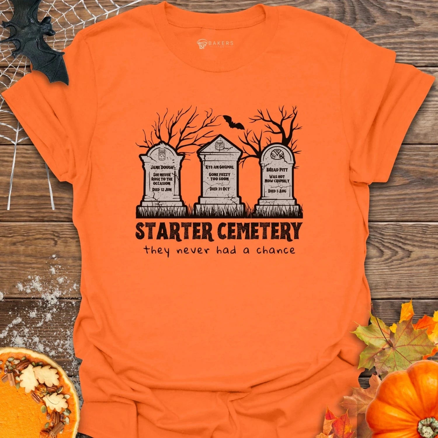 Orange t-shirt with Halloween-themed design featuring gravestones and text on a wooden background.