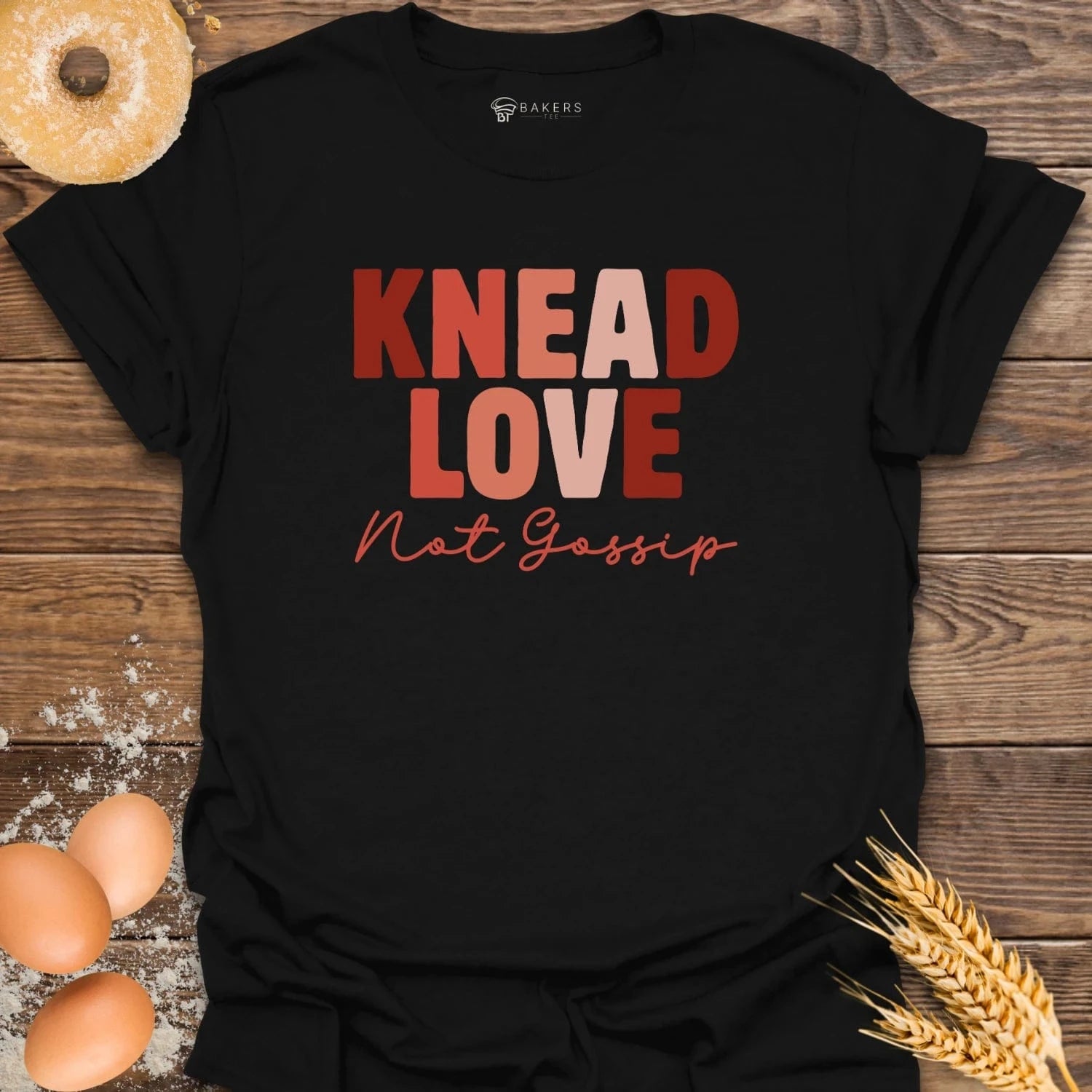 Black t-shirt with 'KNEAD LOVE Not Gossip' text on a wooden surface with eggs and wheat.