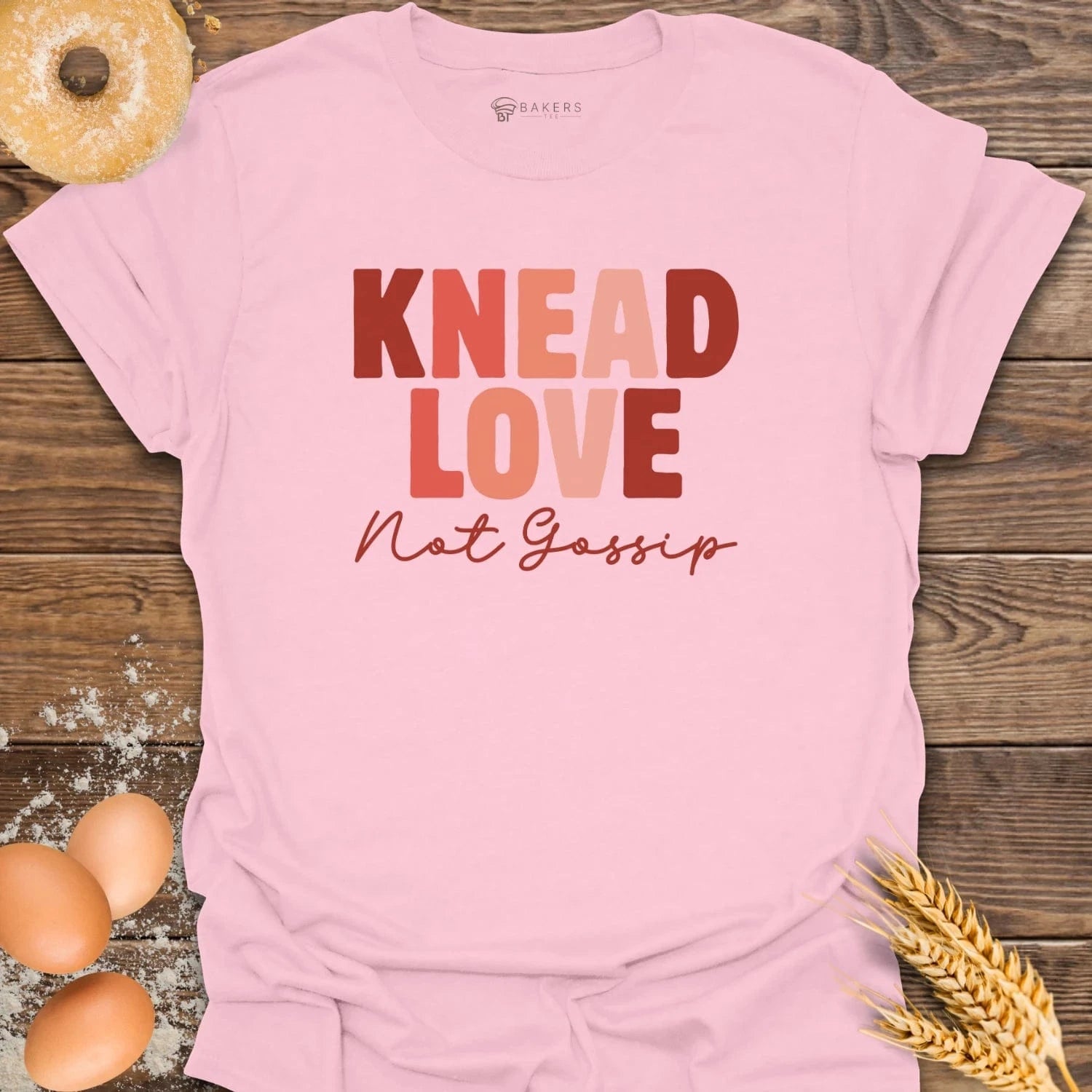 Pink t-shirt with 'Knead Love Not Gossip' text on a wooden surface with eggs and wheat.