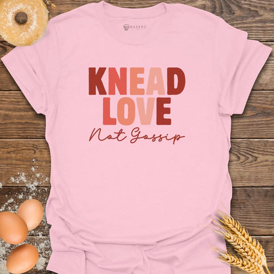 Pink t-shirt with 'Knead Love Not Gossip' text on a wooden surface with eggs and wheat.