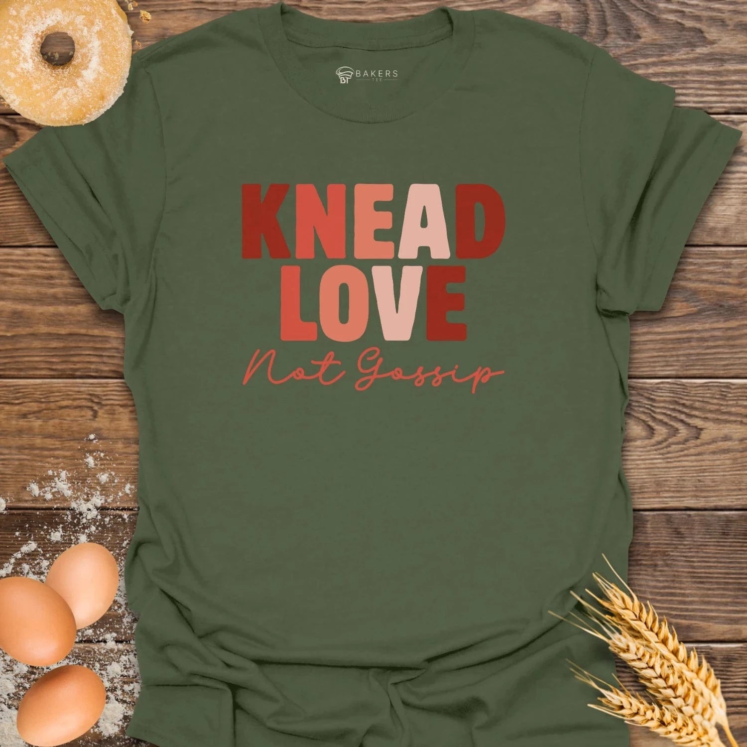 Green t-shirt with 'KNEAD LOVE Not Gossip' text on a wooden surface with bread and eggs.