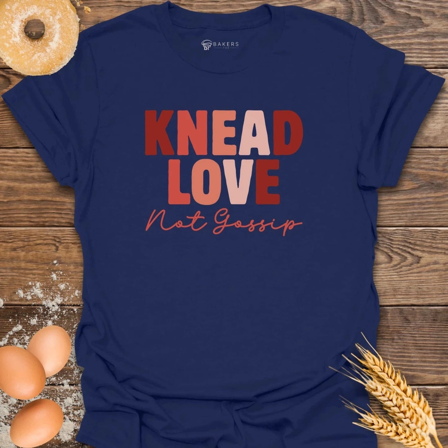 Navy blue t-shirt with 'KNEAD LOVE Not Gossip' text on a wooden surface with eggs and wheat.