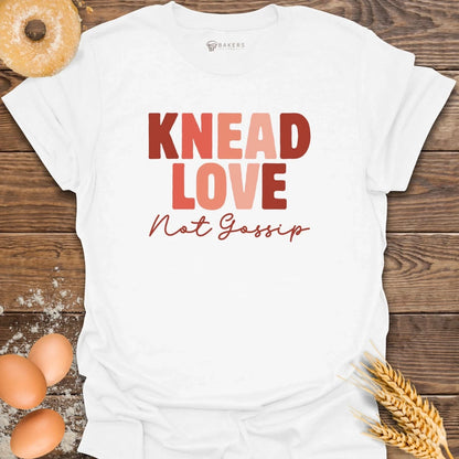 White t-shirt with 'Knead Love Not Gossip' text on a wooden surface with eggs and wheat.
