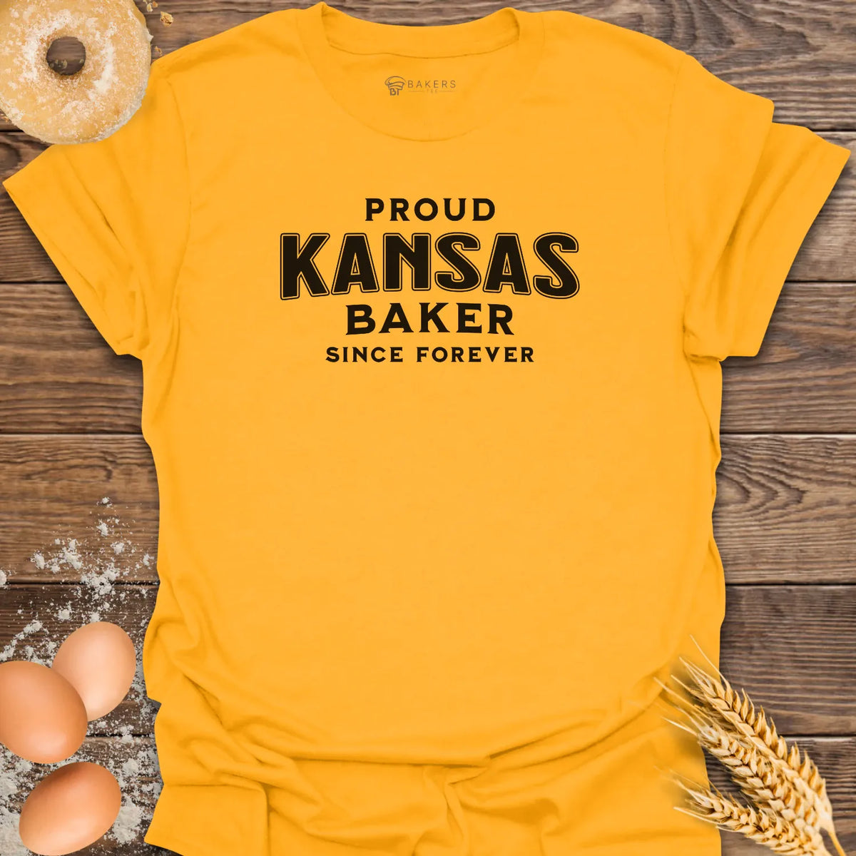 A bright Gold yellow graphic t-shirt laid flat on a rustic wooden background, surrounded by baking-themed props including a donut, eggs, flour, and wheat. The shirt features the text 'PROUD KANSAS BAKER SINCE FOREVER' in black.