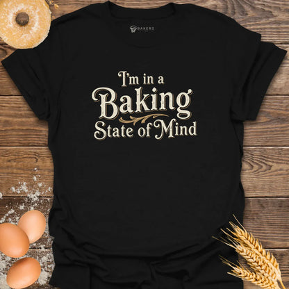 A Black unisex t-shirt laid flat on a wooden surface with eggs, flour, a donut, and wheat beside it. The t-shirt features a Text design reading “I’m in a Baking State of Mind” in blue serif lettering with gold flourishes, styled in a vintage bakery aesthetic