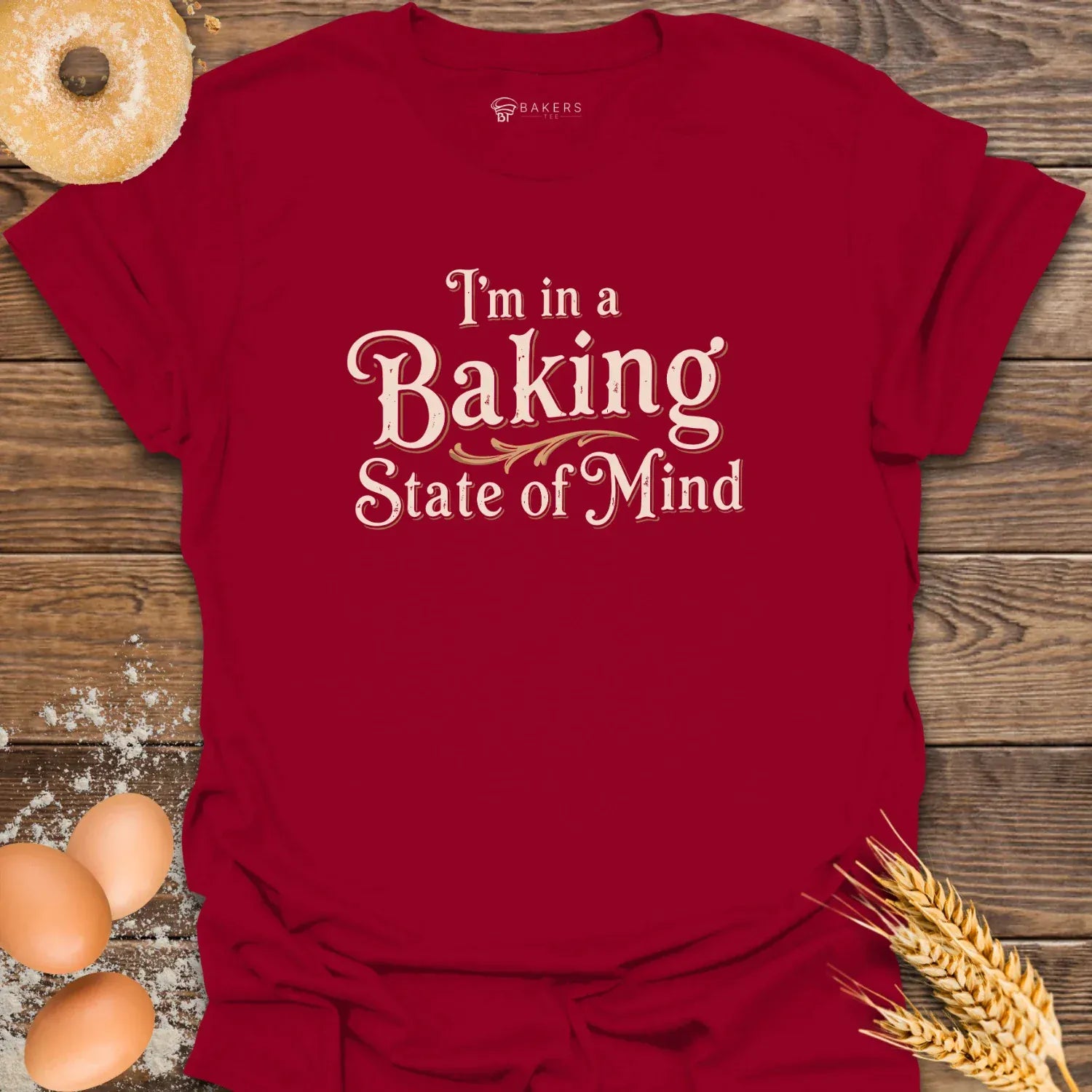 A Cardinal Red unisex t-shirt laid flat on a wooden surface with eggs, flour, a donut, and wheat beside it. The t-shirt features a Text design reading “I’m in a Baking State of Mind” in blue serif lettering with gold flourishes, styled in a vintage bakery aesthetic