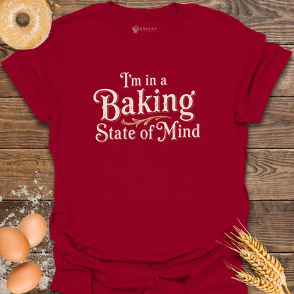 A Cardinal Red unisex t-shirt laid flat on a wooden surface with eggs, flour, a donut, and wheat beside it. The t-shirt features a Text design reading “I’m in a Baking State of Mind” in blue serif lettering with gold flourishes, styled in a vintage bakery aesthetic