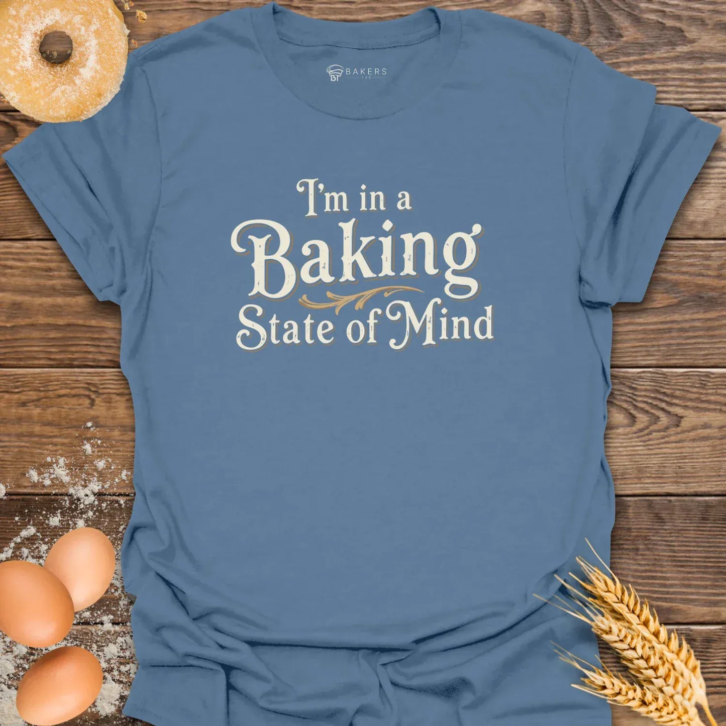 An Indigo Blue unisex t-shirt laid flat on a wooden surface with eggs, flour, a donut, and wheat beside it. The t-shirt features a Text design reading “I’m in a Baking State of Mind” in blue serif lettering with gold flourishes, styled in a vintage bakery aesthetic