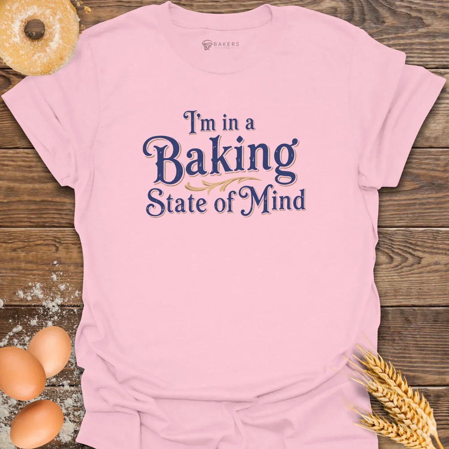 A Light Pink unisex t-shirt laid flat on a wooden surface with eggs, flour, a donut, and wheat beside it. The t-shirt features a Text design reading “I’m in a Baking State of Mind” in blue serif lettering with gold flourishes, styled in a vintage bakery aesthetic