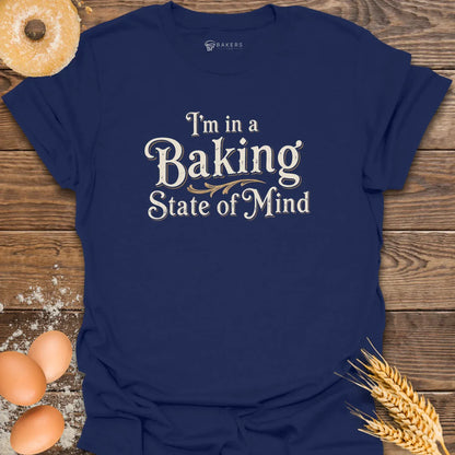 A Navy unisex t-shirt laid flat on a wooden surface with eggs, flour, a donut, and wheat beside it. The t-shirt features a Text design reading “I’m in a Baking State of Mind” in blue serif lettering with gold flourishes, styled in a vintage bakery aesthetic