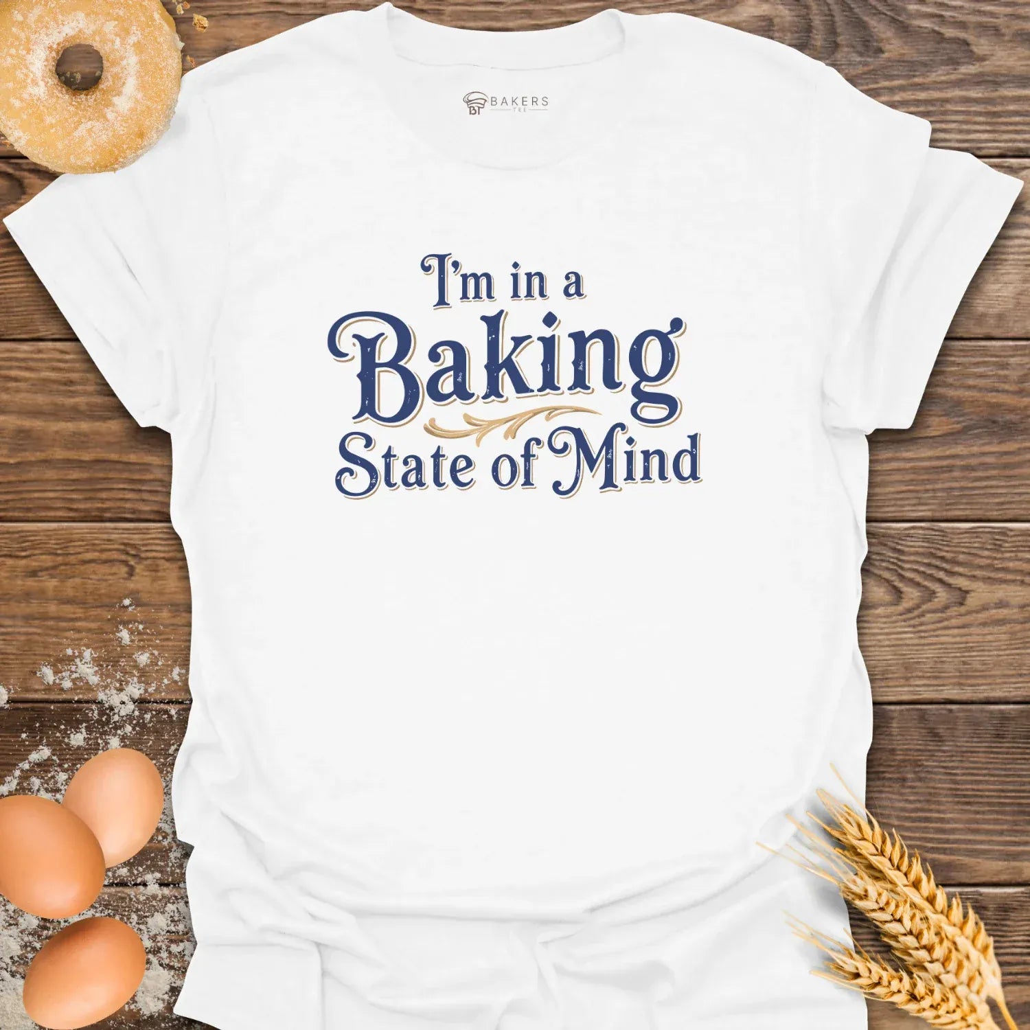 A white unisex t-shirt laid flat on a wooden surface with eggs, flour, a donut, and wheat beside it. The t-shirt features a Text design reading “I’m in a Baking State of Mind” in blue serif lettering with gold flourishes, styled in a vintage bakery aesthetic
